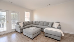 Ein Wohnzimmer mit einem grauen Ecksofa, zwei Poufs und einer weißen Decke.