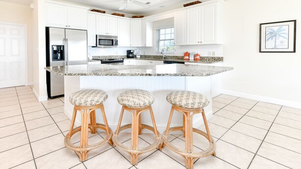Eine Küche mit weißen Schränken, einem Edelstahl-Kühlschrank, einer Mikrowelle und einer Kaffeemaschine. Drei Holz-Barke stools mit gemusterten Kissen sind vorhanden.