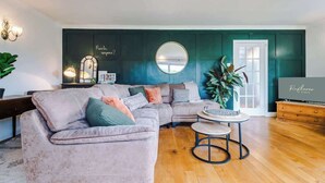 Een woonkamer met een hoekbank, een ronde spiegel, een klein rond tafeltje en een televisie.