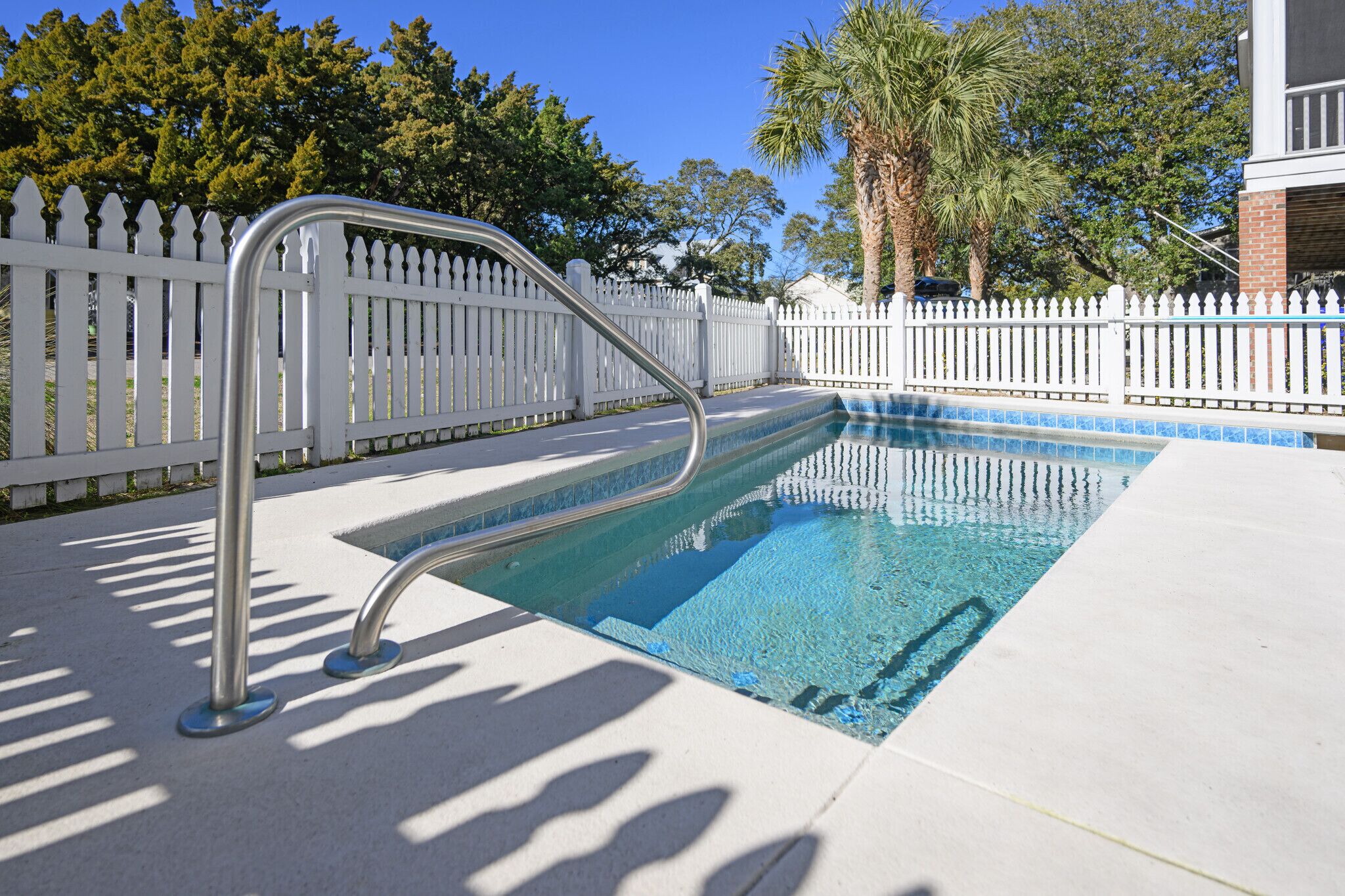 A swimming pool with a metal ladder, surrounded by a white picket fence and palm trees.