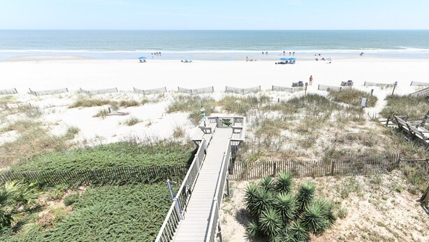 Een strand met een houten pier die naar het strand leidt, zandduinen en schaarse begroeiing.