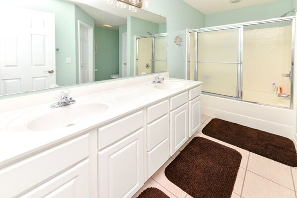 A bathroom with two sinks, a large mirror, a shower, and brown rugs on the floor.