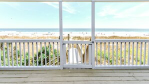 Blick auf den Strand von einer überdachten Veranda mit weißer Brüstung, Holzterrasse und einer Glastür zum Strand.