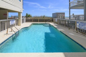 Une piscine rectangulaire sans sauveteur, entourée d’une clôture en bois, et d’où l’on a une vue dégagée sur la plage en arrière-plan.