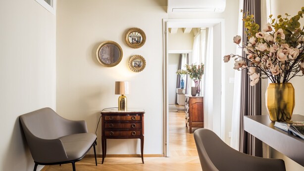 A modern living room with a wooden herringbone floor, a grey sofa, a small wooden side table with a lamp, and a vase with flowers on a console table.
