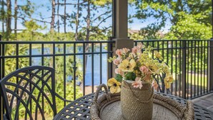 A wicker table with a woven placemat, a wicker chair, and a vase of flowers on a balcony with a metal railing.