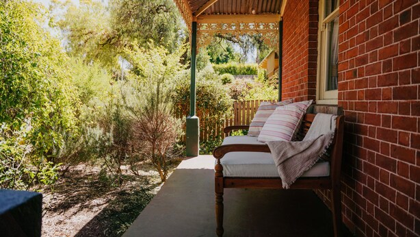 A covered outdoor seating area with a wooden bench and cushions, surrounded by greenery and a brick wall.