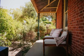 A covered outdoor seating area with a wooden bench and cushions, surrounded by greenery and a brick wall. - The Redbrick House (Castlemaine)