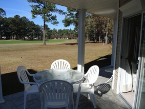 A covered patio with a glass table and white plastic chairs, overlooking a golf course. - 2301 2 Bedroom Villa by RedAwning (Calabash)