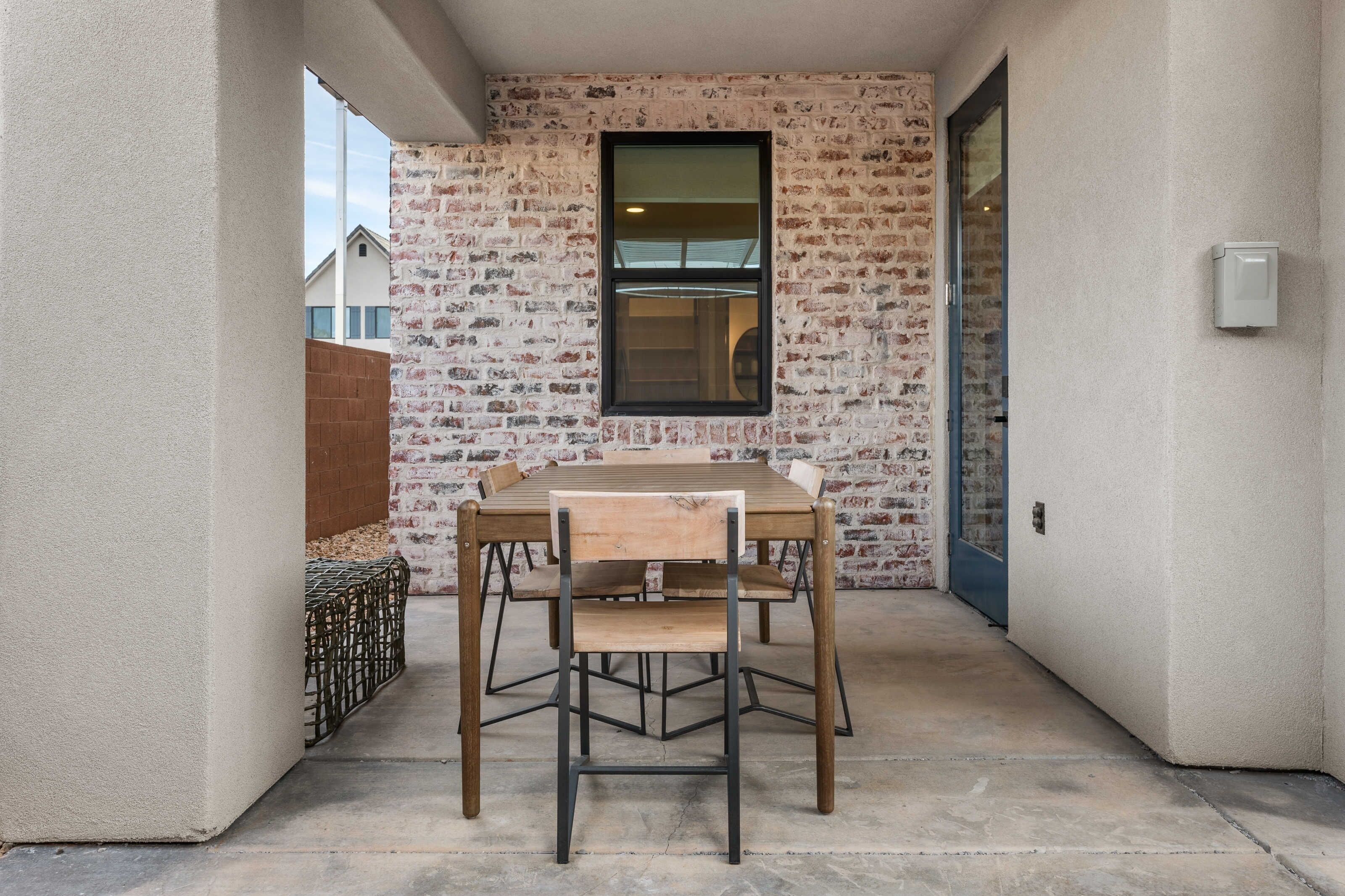 A small patio with a wooden table and chairs, a brick wall, and a window.