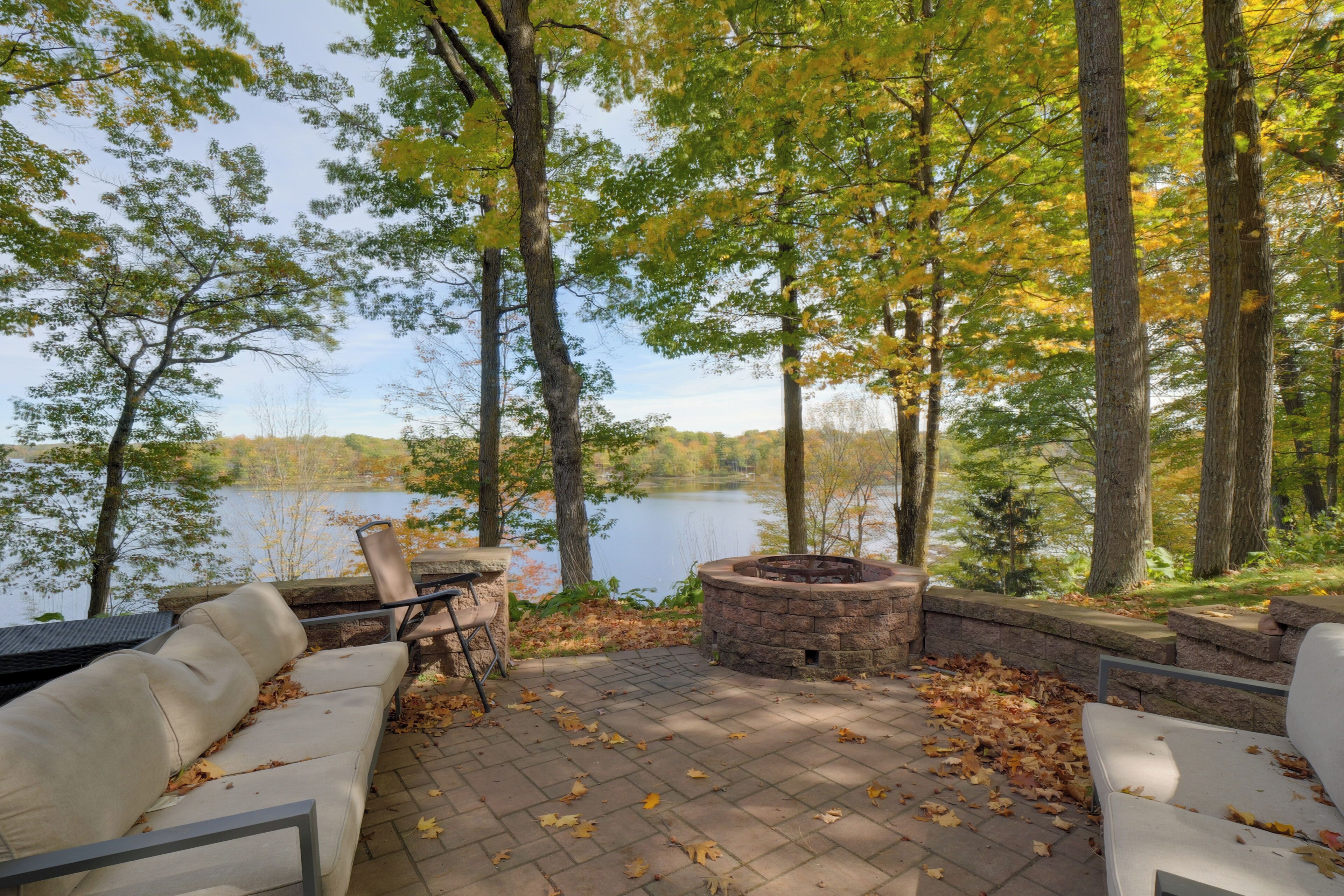 Un espace extérieur aménagé avec des sièges, un foyer en pierre et une vue sur un lac entouré d’arbres.