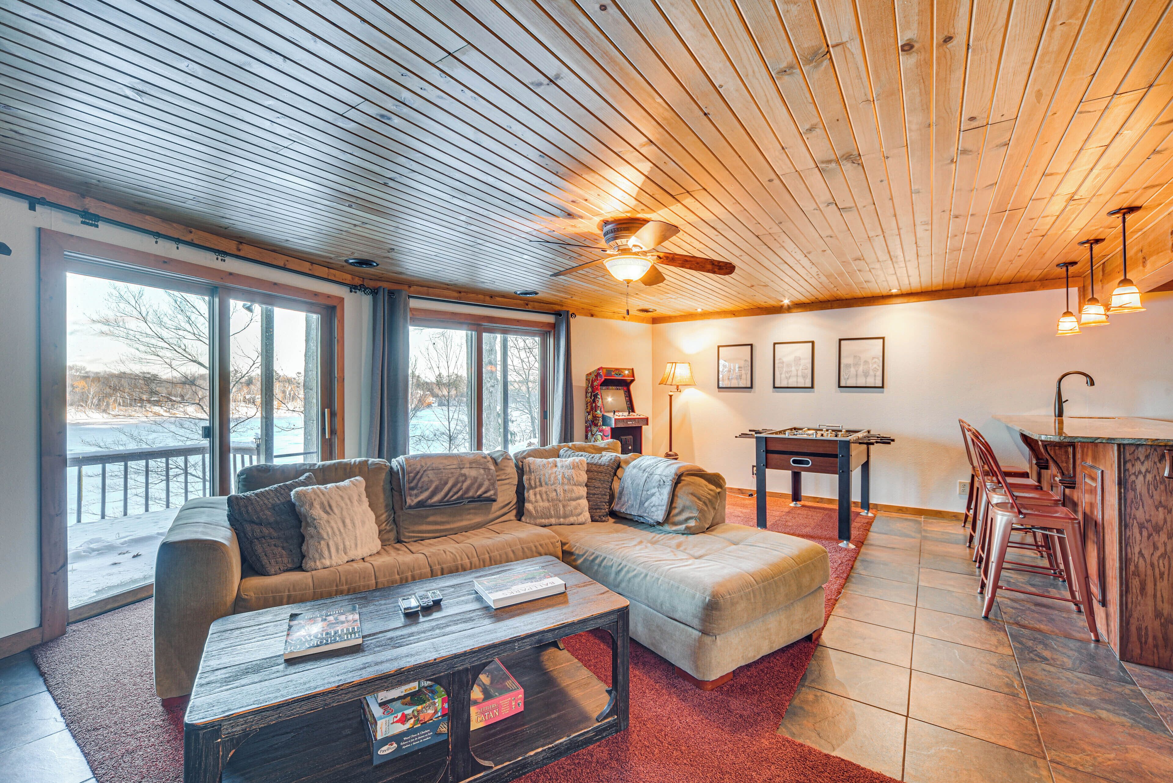 A spacious living room with a large sectional sofa, a coffee table, and a pool table. There are large windows providing a view of the outdoors.
