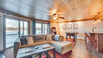 A spacious living room with a large sectional sofa, a coffee table, and a pool table. There are large windows providing a view of the outdoors.