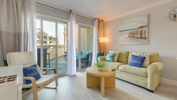 Un salon comprenant un canapé, un fauteuil et une table basse, une porte-fenêtre coulissante donnant sur un balcon, et un tableau représentant une scène de plage.