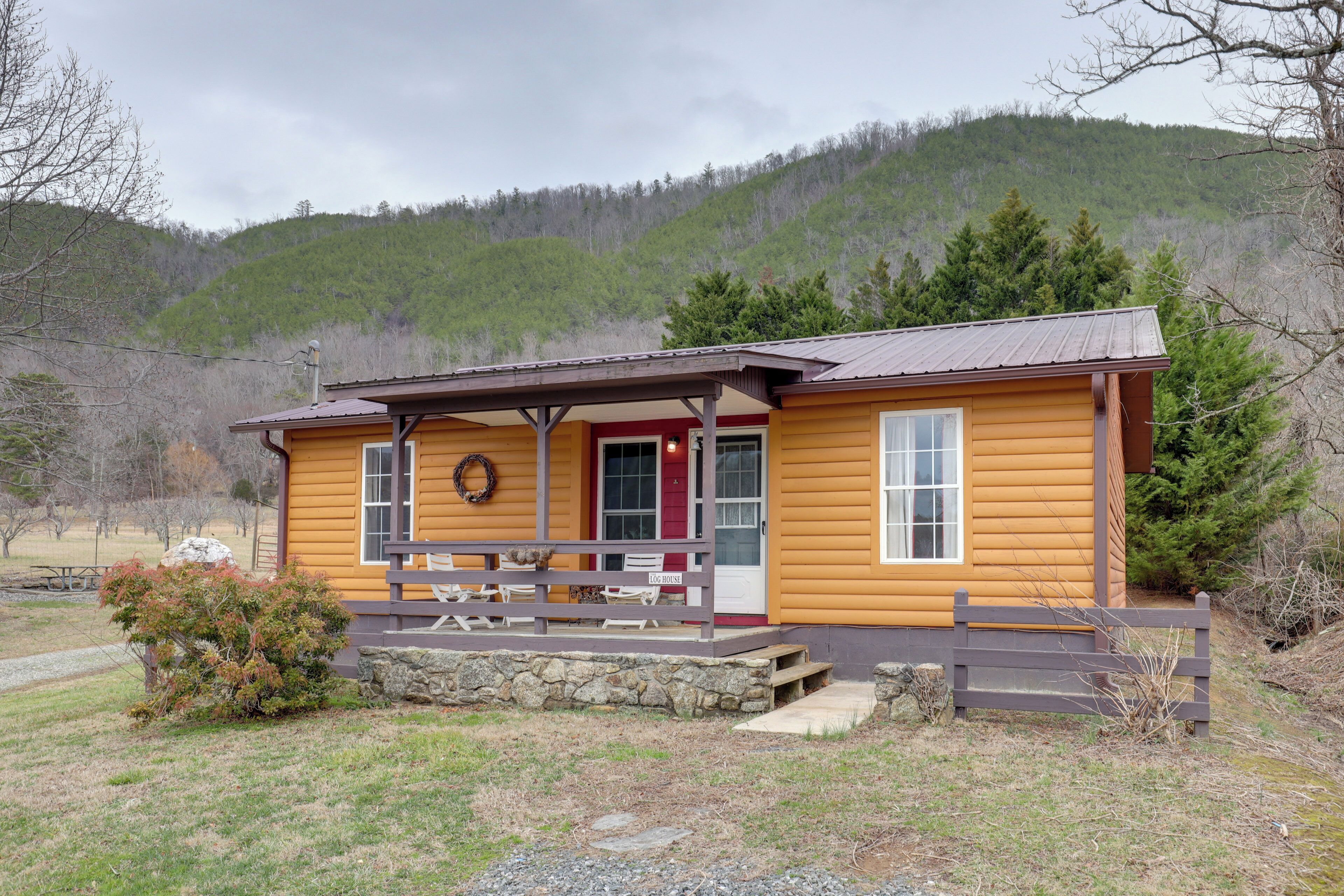 A wooden cabin with a porch, stone foundation, and a view of a forested hill.