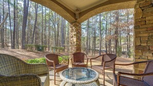 A covered outdoor seating area with wicker furniture and a glass table, surrounded by a stone wall and overlooking a forest. - Chic Greensboro Condo w/ Lake Oconee Access! (Greensboro)