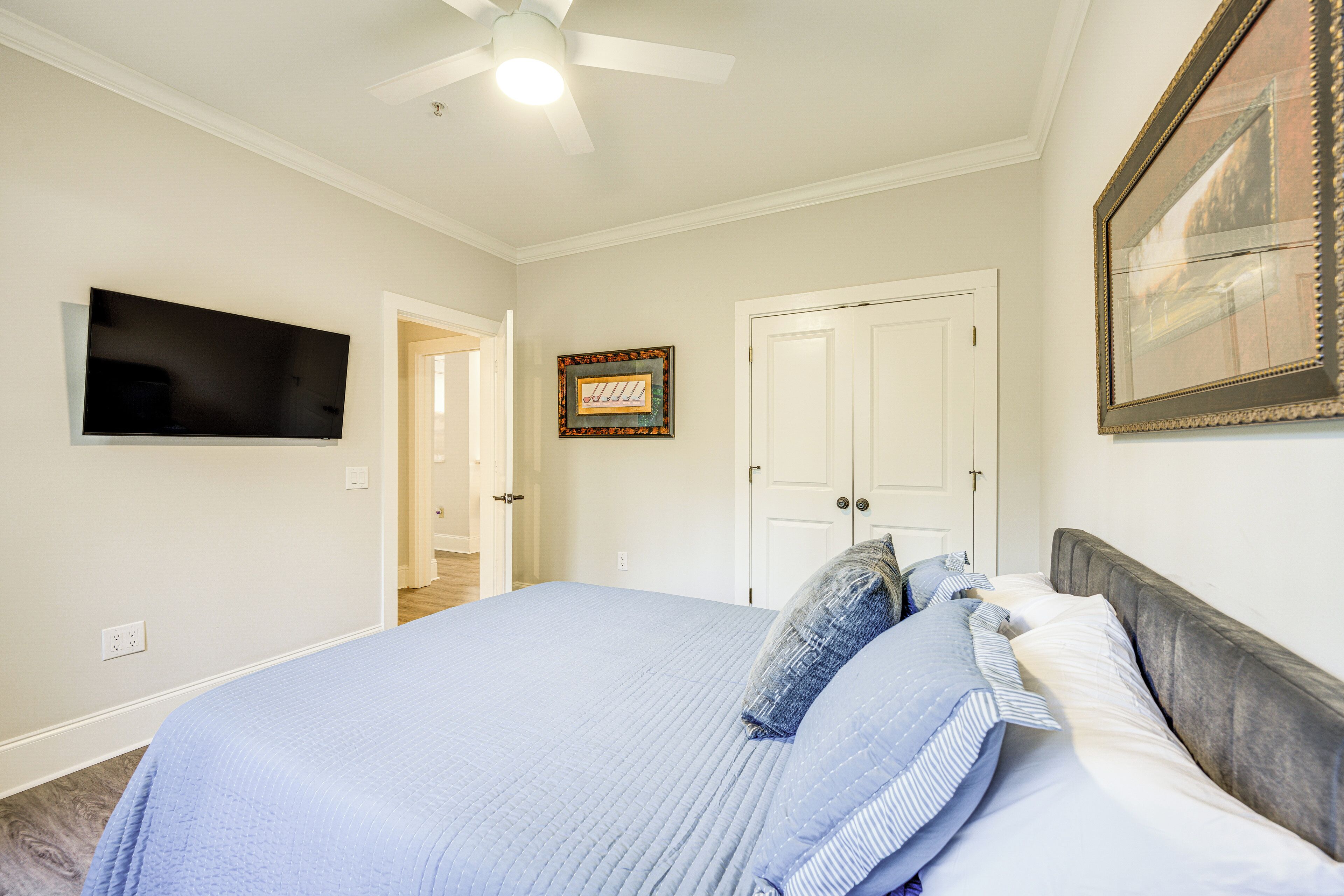 A bedroom with a bed, a ceiling fan, a TV mounted on the wall, and a door leading to another room.
