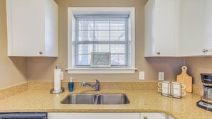 A kitchen with a granite countertop, stainless steel sink, and white cabinets. There is a window with blinds, a coffee maker, and two coffee mugs on the counter. - Swim, Golf & Unwind: Barefoot Resort Condo (North Myrtle Beach)