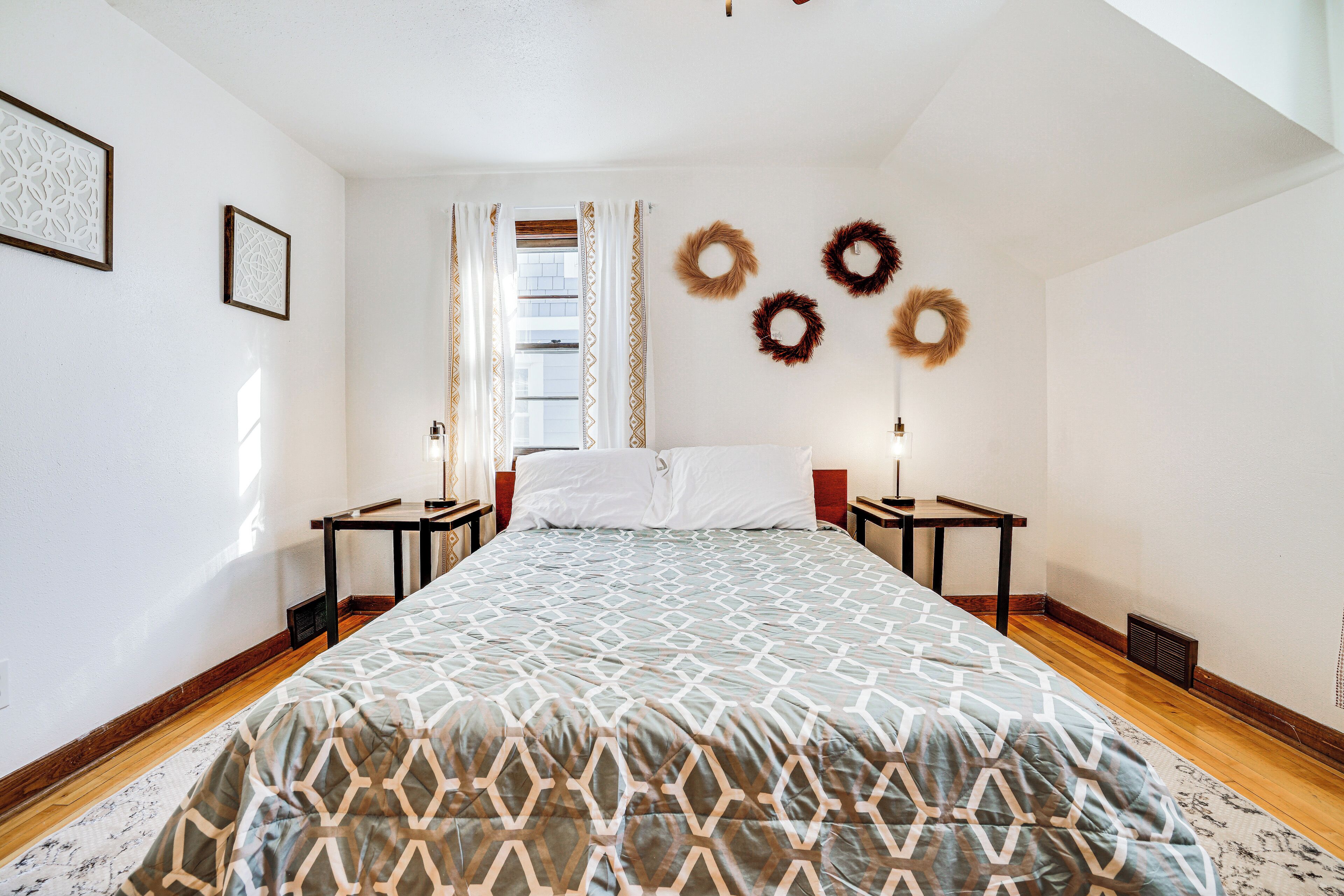 A bedroom with a large bed, two bedside tables, a window with curtains, and three wall-mounted wreaths.