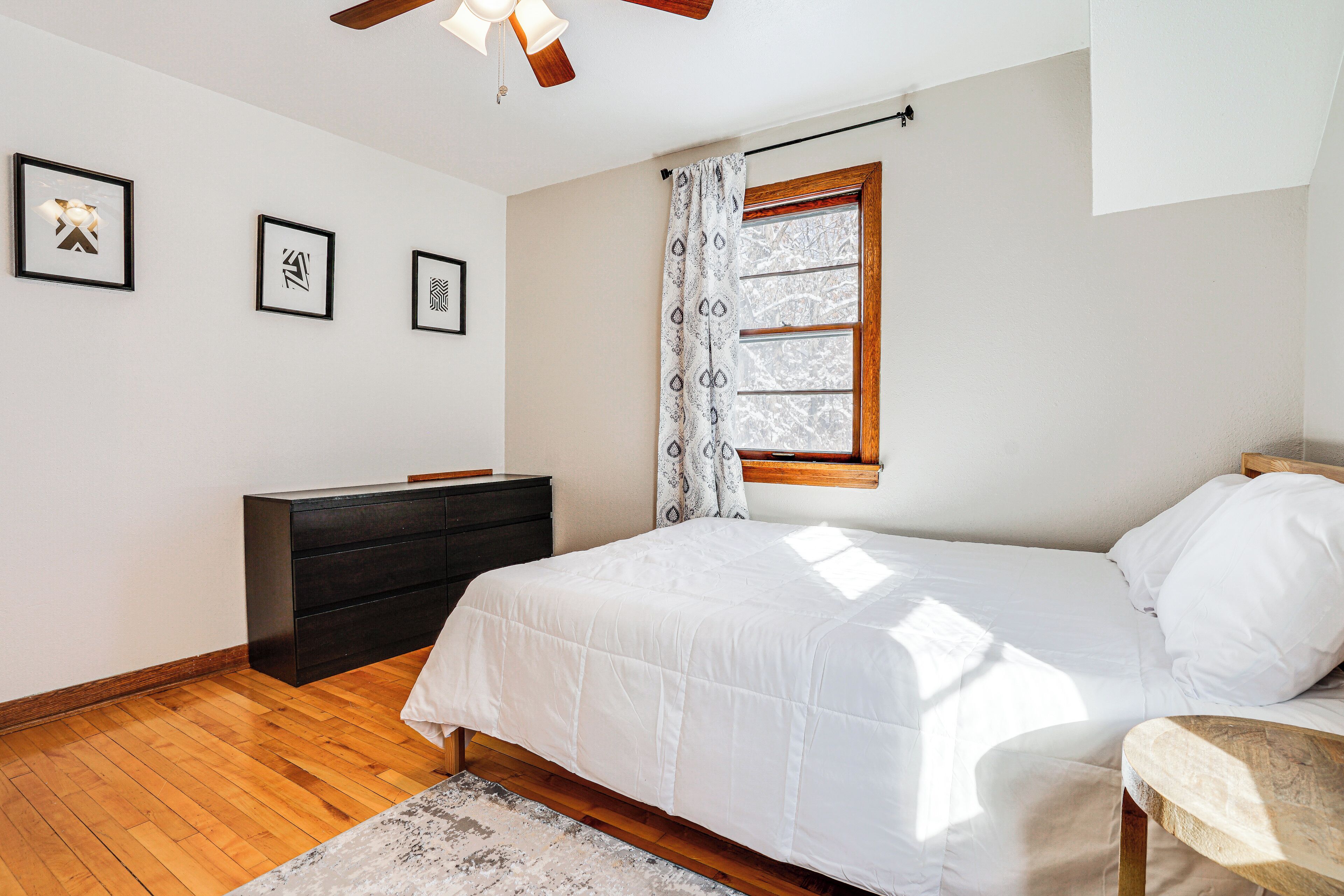 Un dormitorio con cama, cómoda, ventilador de techo y ventana con cortinas.
