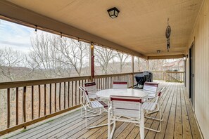 A covered outdoor patio with a wooden deck, white metal chairs, a round table, and a barbecue grill. - Spacious Holiday Island Escape w/ Deck & Gas Grill (Eureka Springs)
