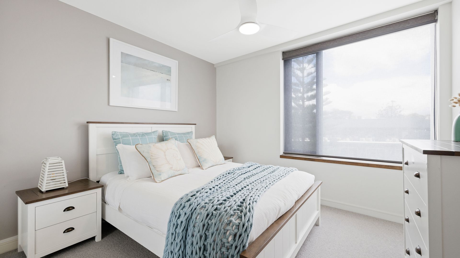 A bedroom with a bed, nightstand, dresser, and a large window with a view.