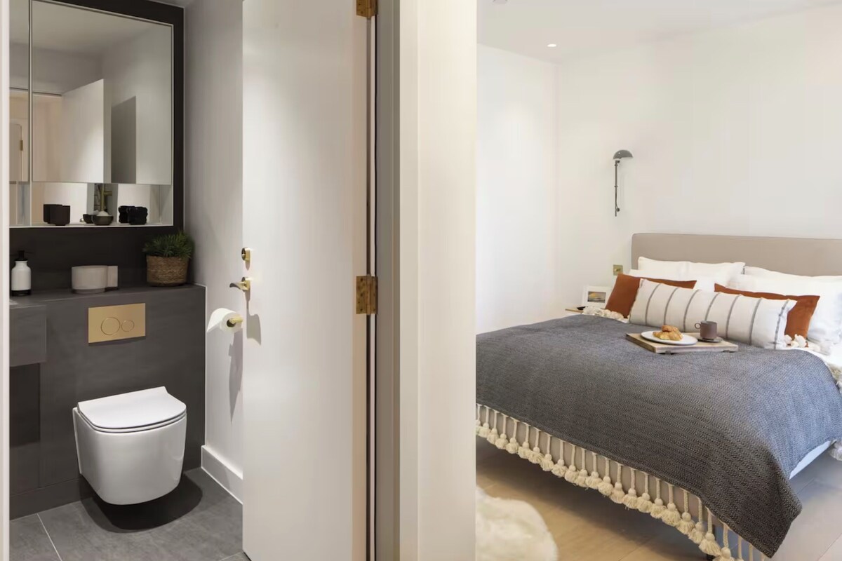 A modern bathroom with a toilet, sink, and mirror. The bedroom features a bed with a gray blanket and white pillows.