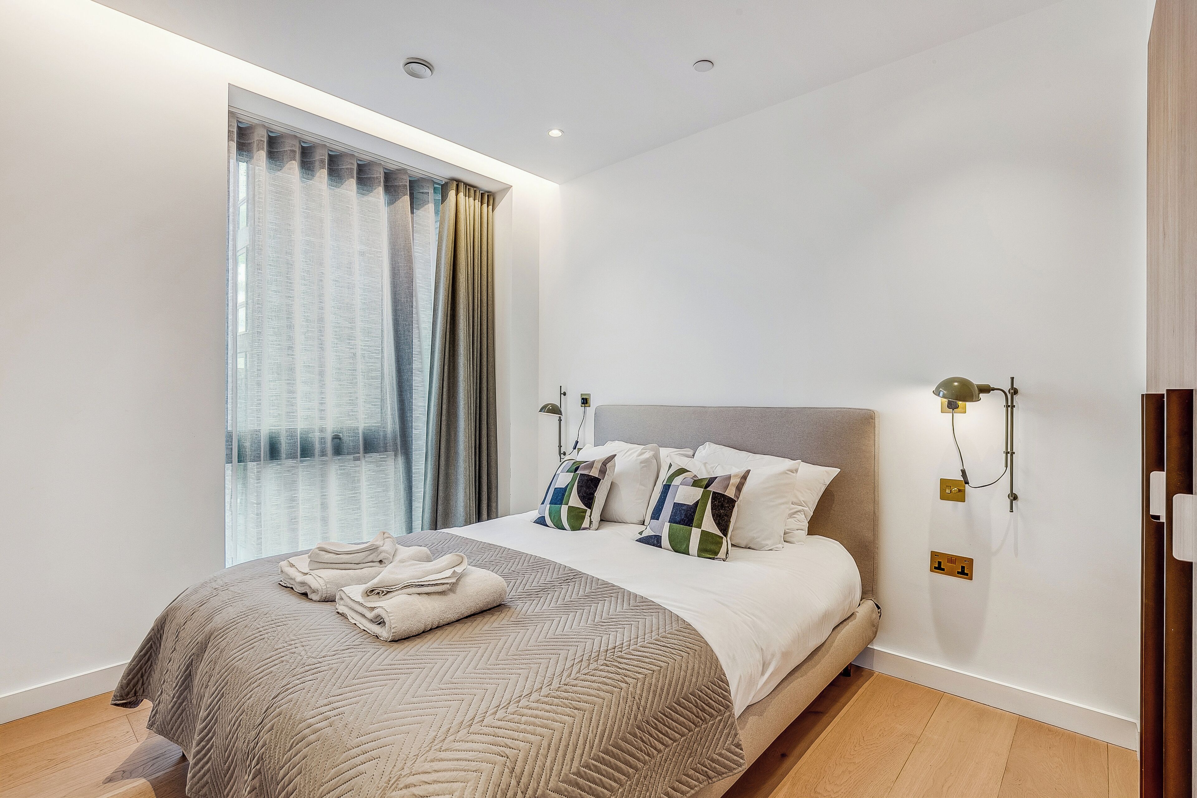 Una cama bien hecha con una colcha de diseño greca, un par de toallas dobladas y almohadones decorativos.