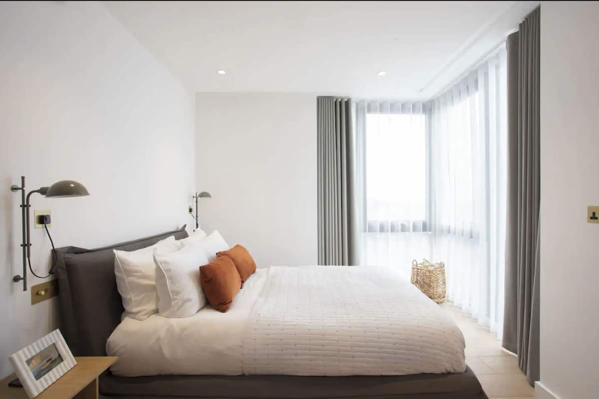 Un dormitorio moderno con una cama grande, dos lámparas de noche, un marco de foto en una mesita de noche y vistas al exterior a través de cortinas translúcidas.