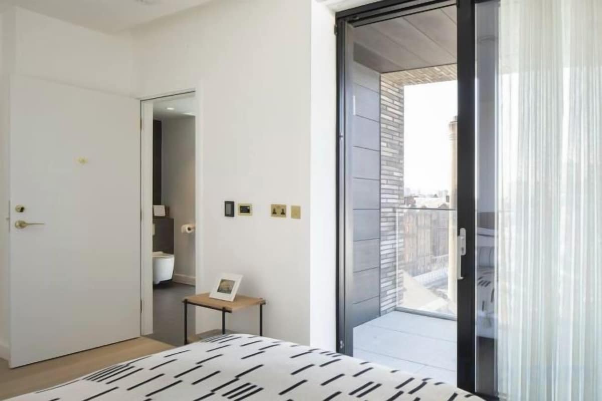 A bedroom with a bed, a door, a small table with a picture, and a view through a glass door.