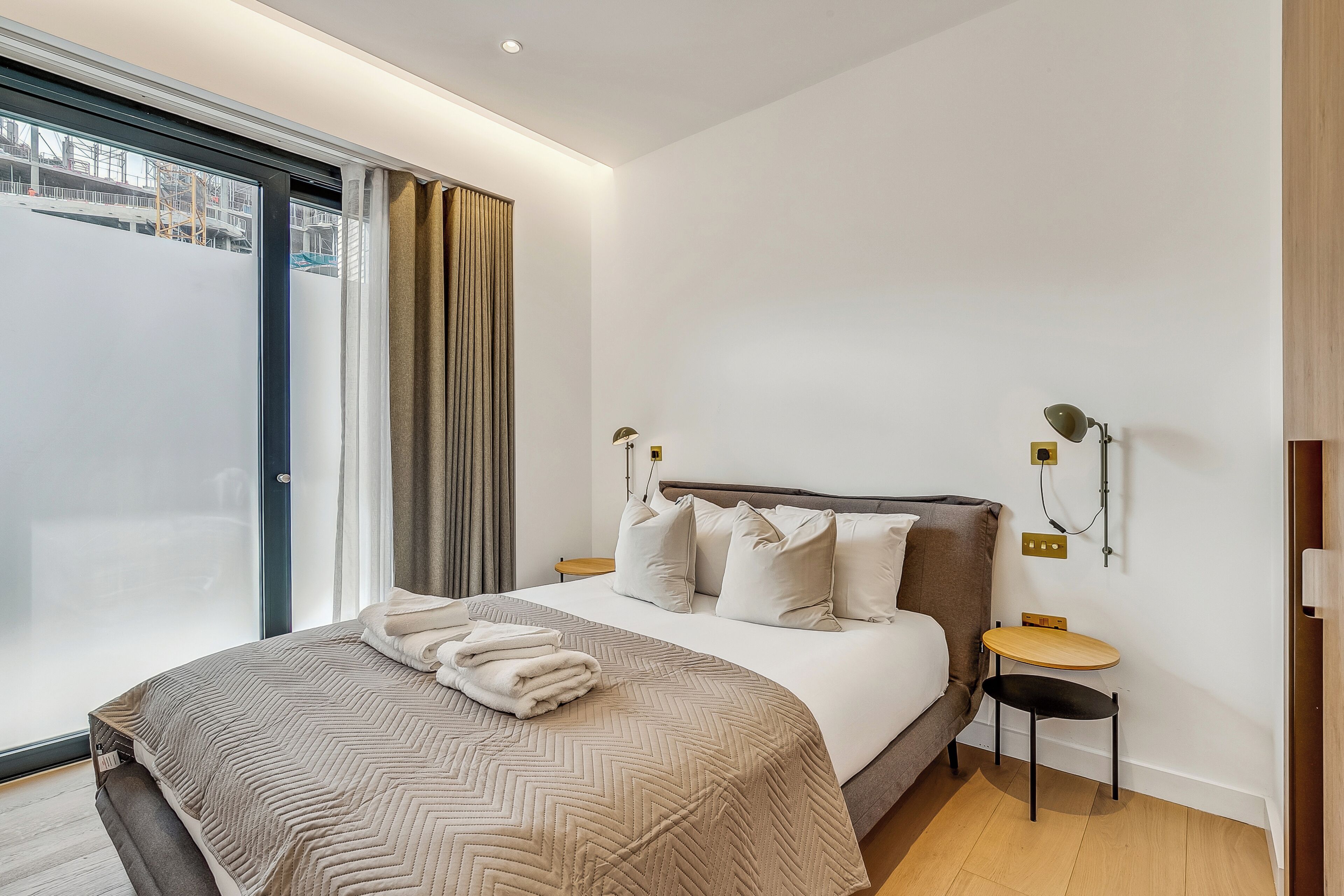 Ein modernes Hotelzimmer mit einem großen Bett, einem kleinen Tisch, einer Wandlampe und Blick durch eine Glastür.