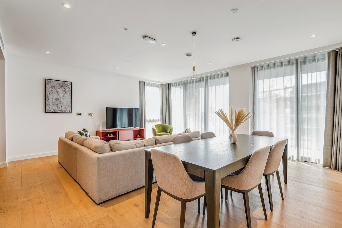 Una sala de estar moderna con un sofá grande, una mesa de comedor y sillas. Hay un televisor y un jarrón decorativo sobre la mesa.