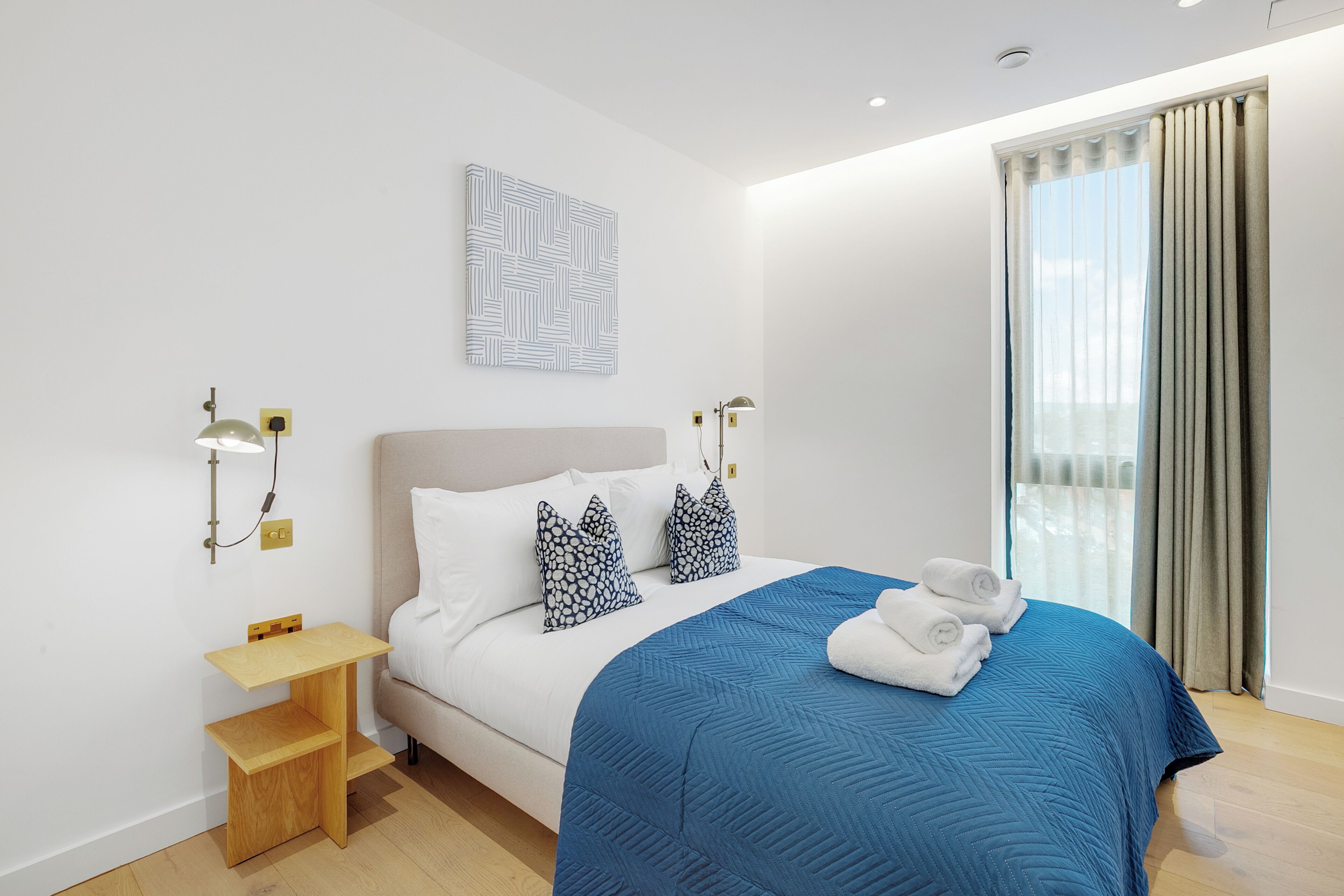 Una habitación de hotel moderna con una cama, una mesita de noche, una lámpara de pared y una ventana con cortinas.