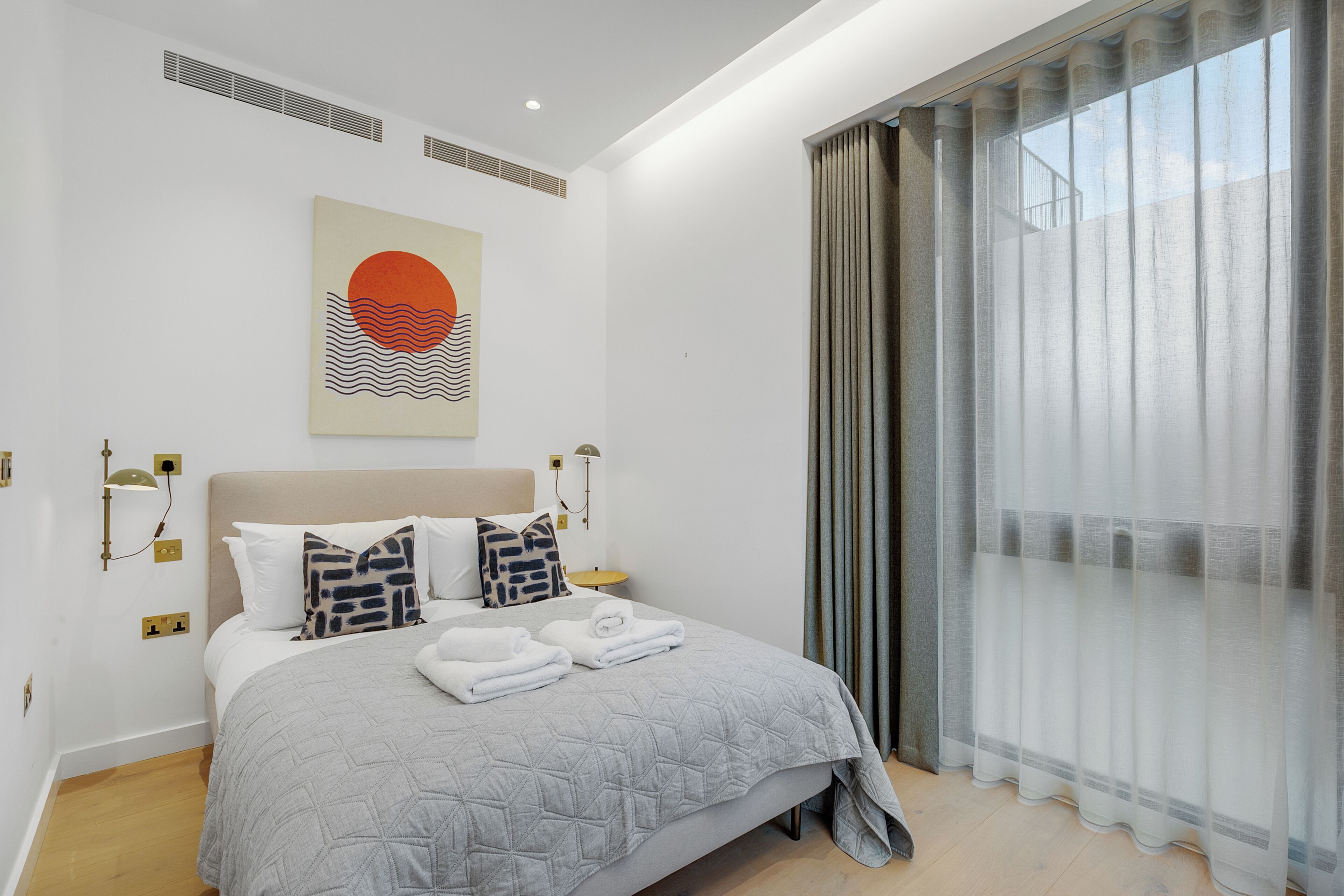 Habitació d'hotel moderna amb un llit gran, una obra penjada a la pared, una finestra amb cortines i una taula de nit amb una làmpada.