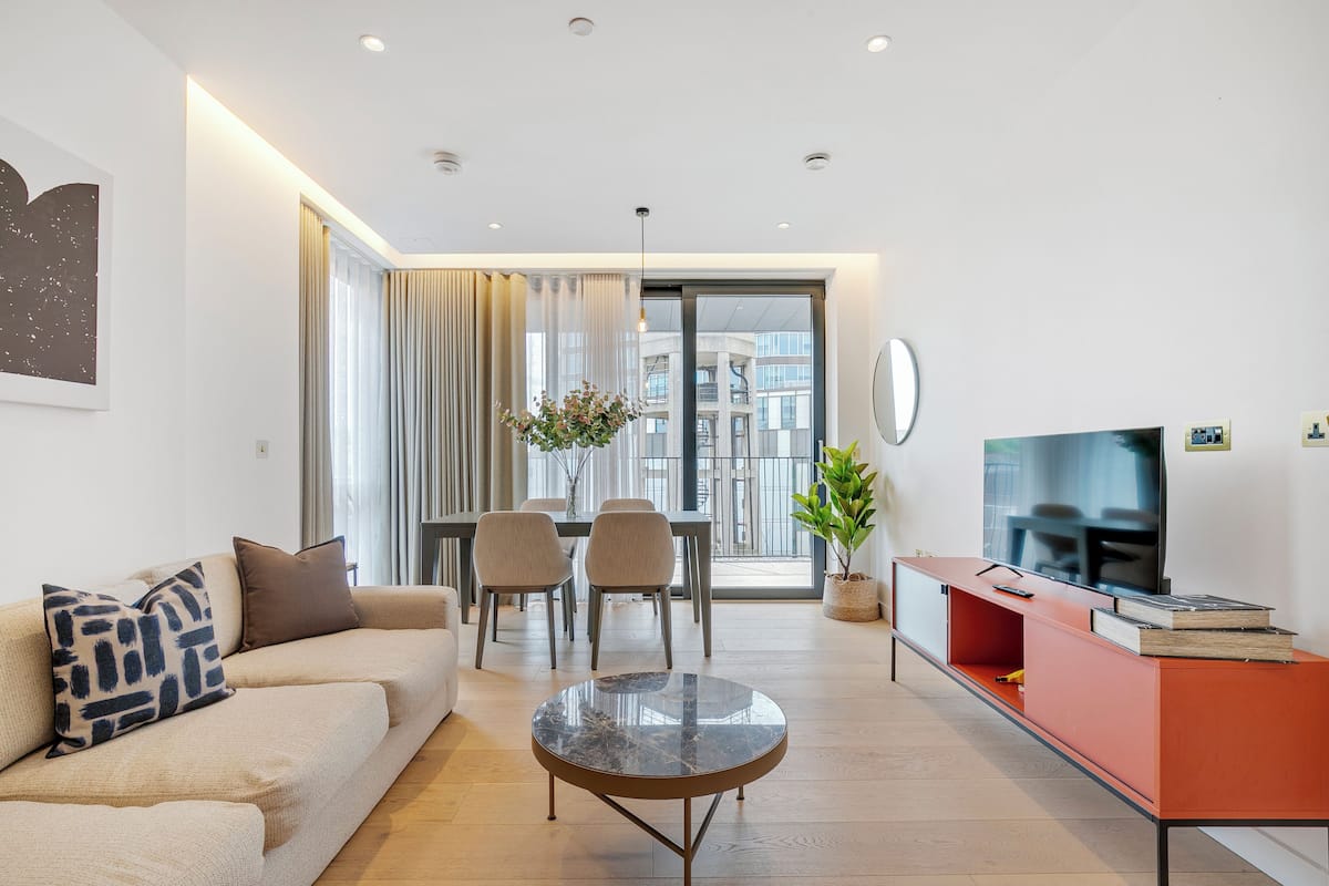 Una sala de estar moderna con sofá, mesa de centro, comedor con sillas, televisor y balcón con vistas a edificios.