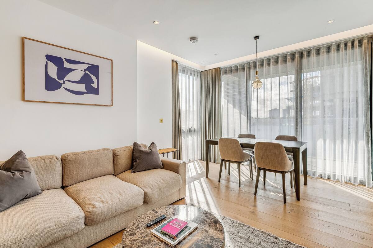 A modern living room with a beige sofa, a coffee table with a book, and a dining area with a table and chairs.