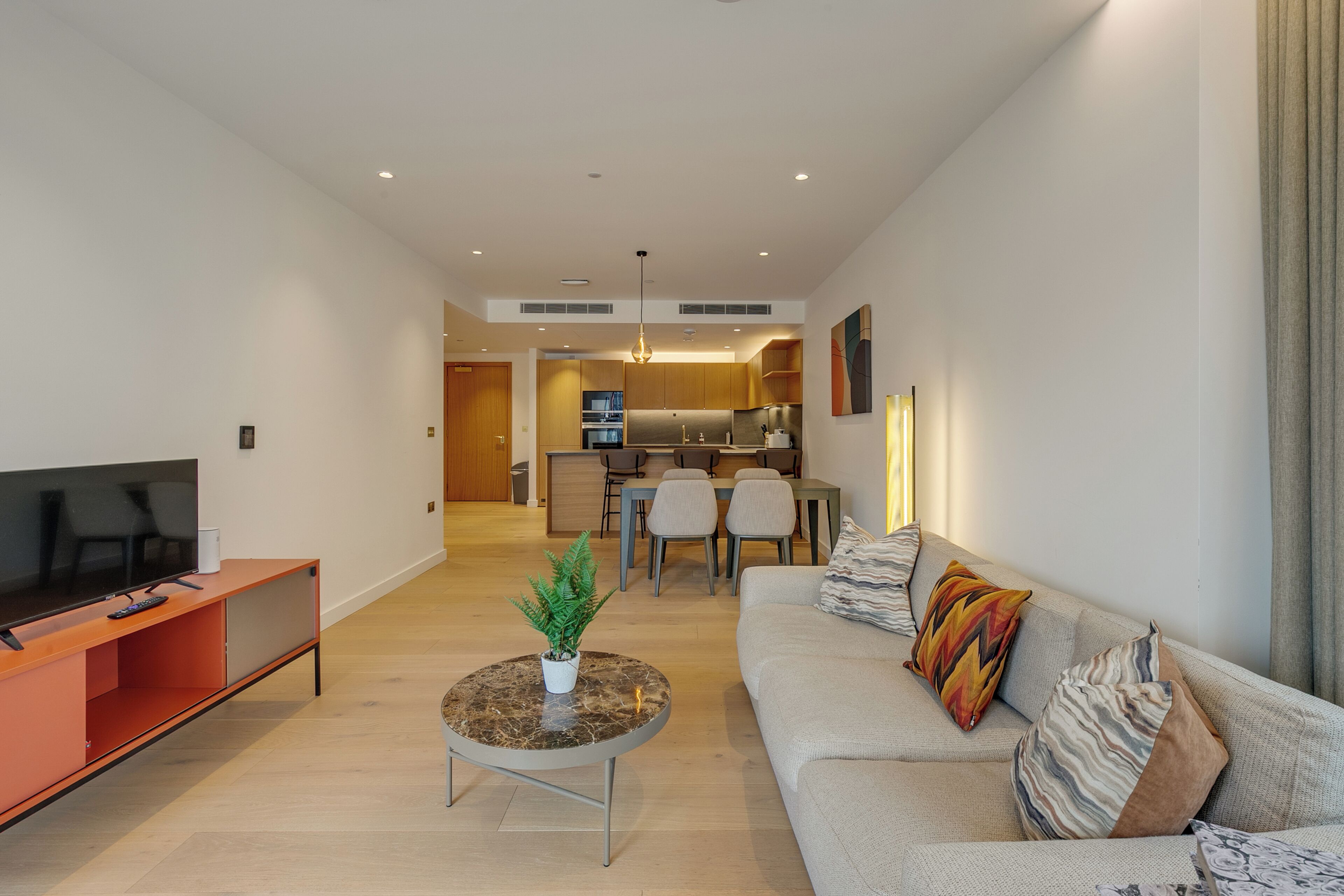 A modern living room with a flat-screen TV, a sofa with patterned cushions, a coffee table with a plant, and a dining area with chairs and a kitchen in the background.