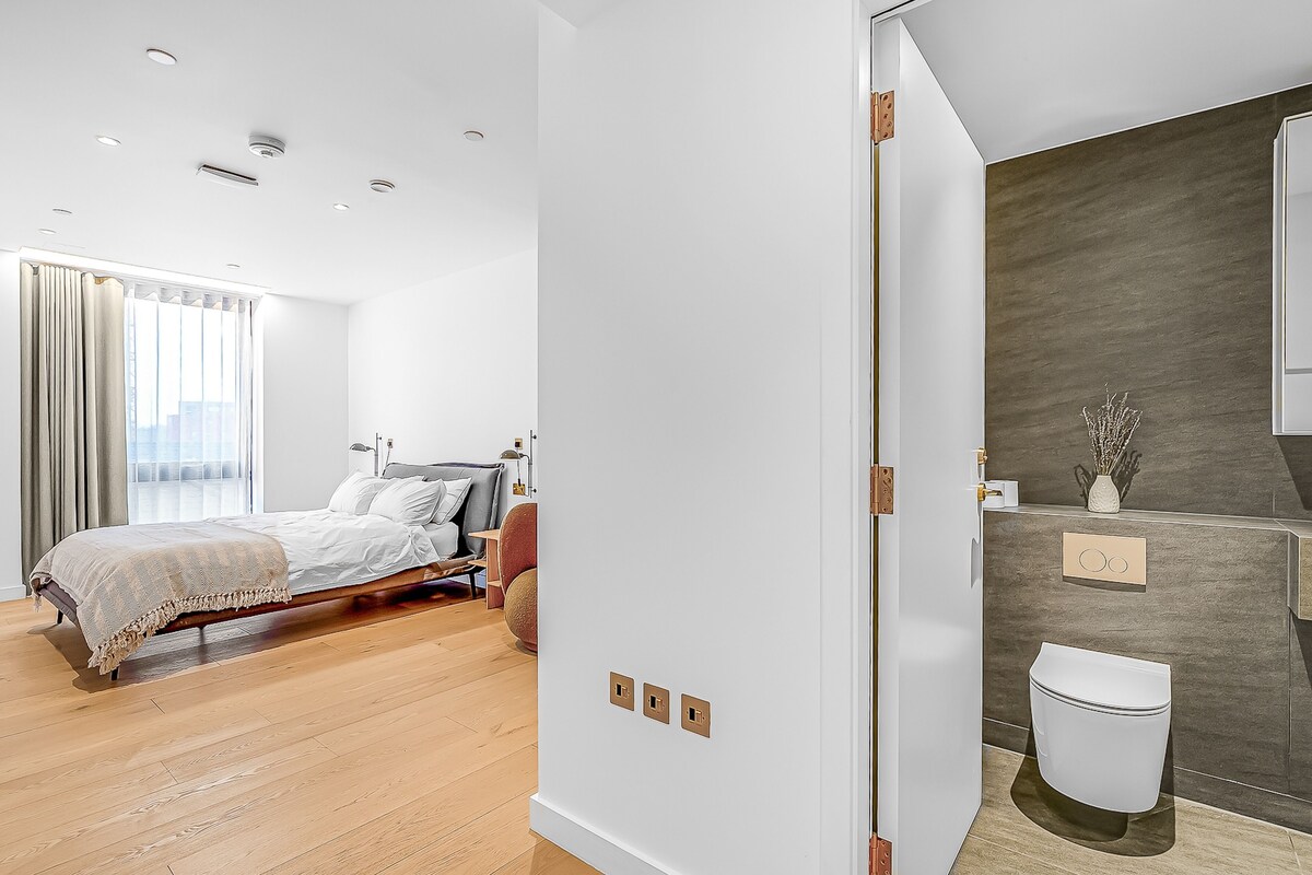 A modern bedroom with a wooden floor, a bed with a beige blanket, a city view through large windows, and a bathroom with a modern design.