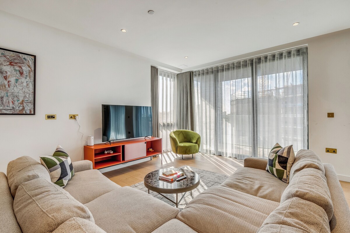 A modern living room with a large sectional sofa, a round coffee table, a red entertainment unit, a green armchair, and a large window with sheer curtains.