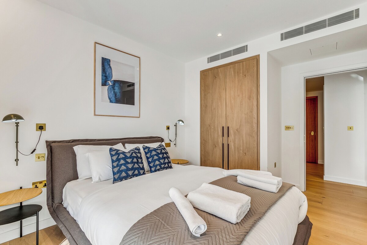 A neatly arranged hotel room with a large bed, a wooden wardrobe, a framed abstract painting, and a small table with a lamp.