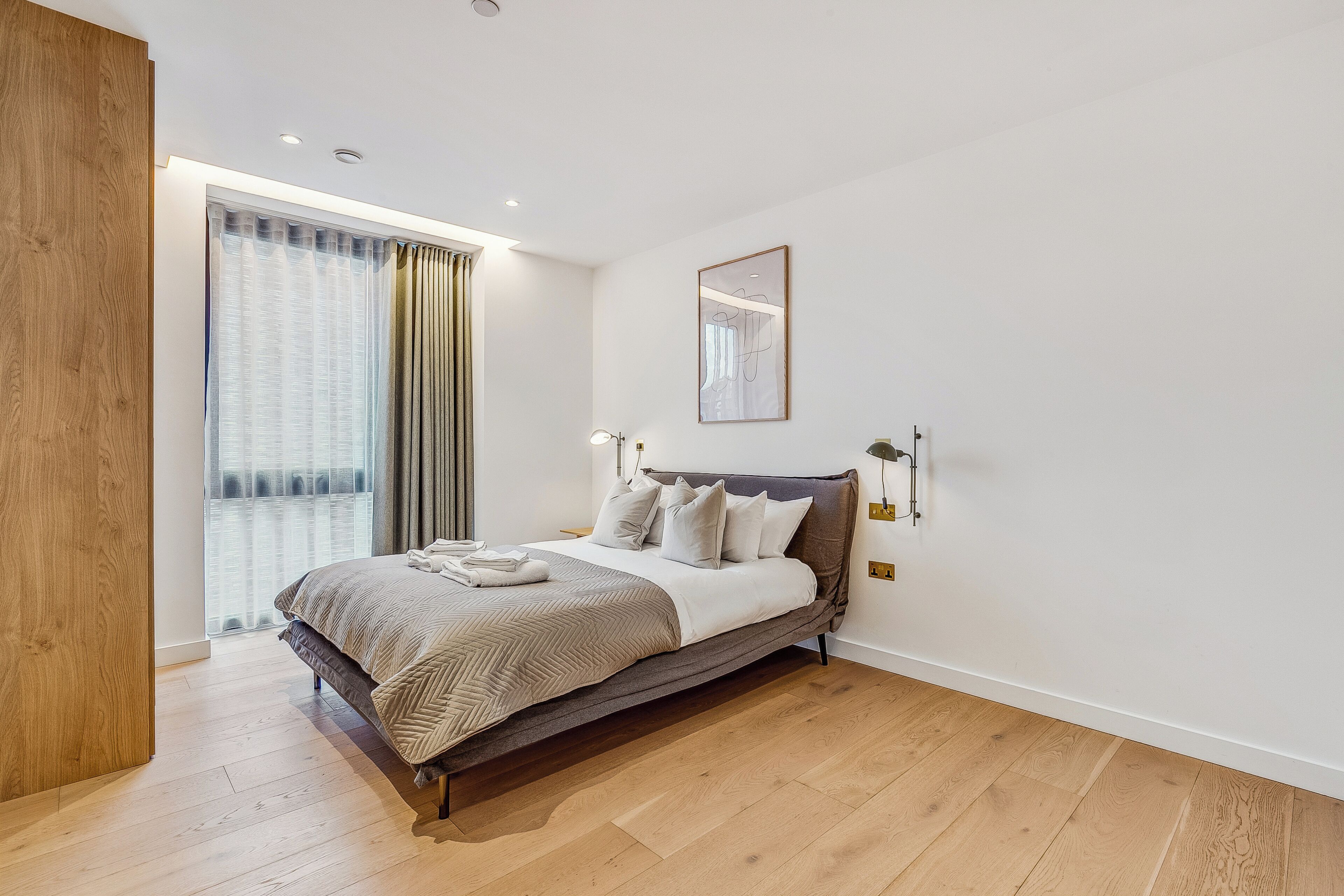 Un dormitorio moderno con una cama grande, piso de madera, una ventana con cortinas y un espejo fijado a la pared.