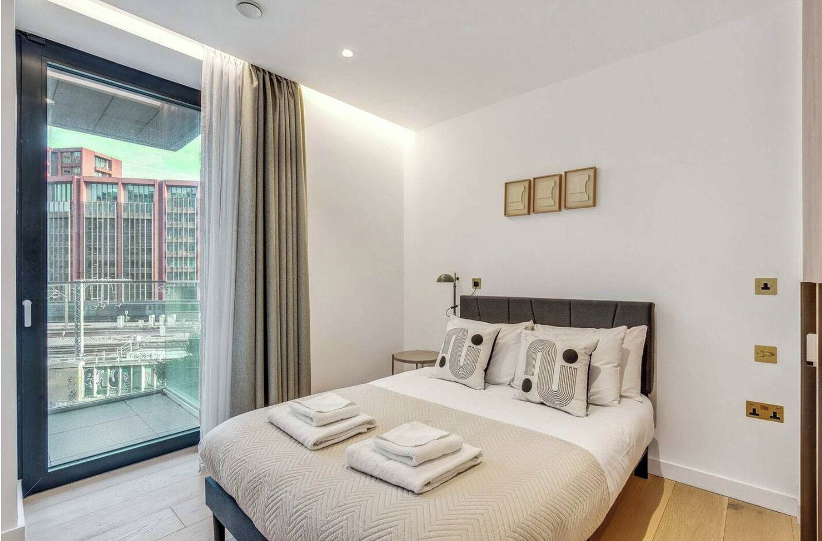 A modern bedroom with a large bed, a view of buildings through a glass door, and a balcony.