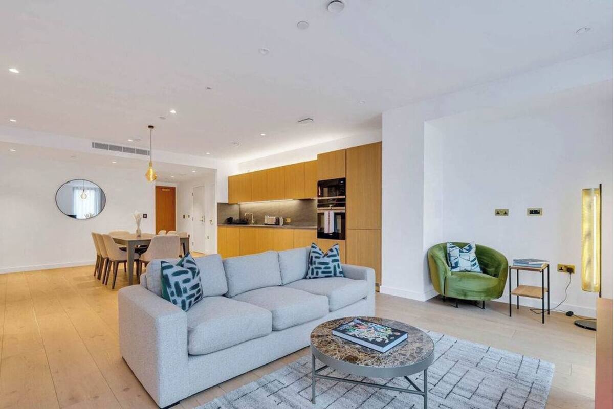 A modern living room with a grey sofa, a round coffee table, and a green armchair. There is a dining area with a round table and chairs, and a kitchen in the background.