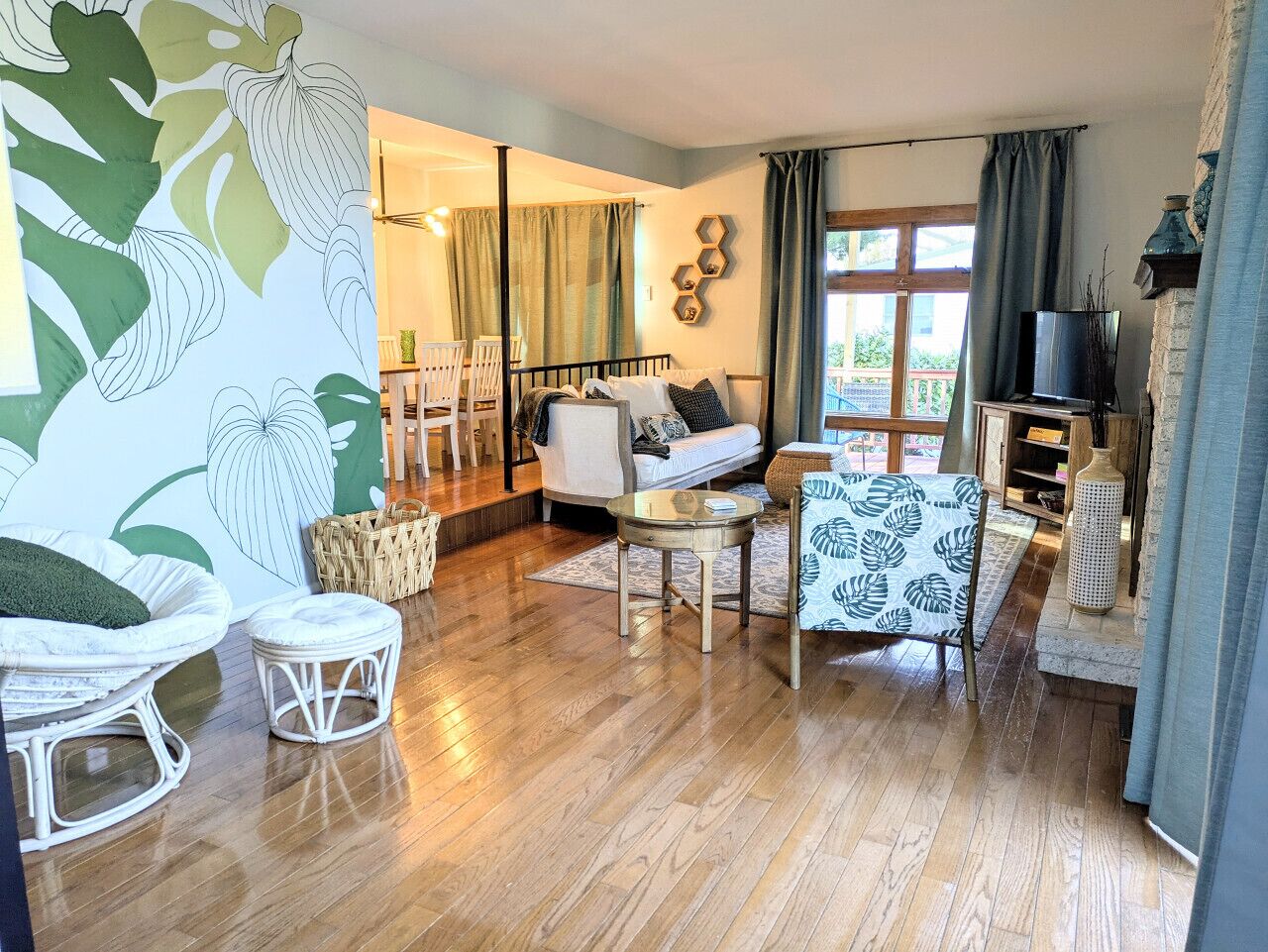 A cozy living room with a sofa, a small round table, and a chair. There is a large window with curtains, a bookshelf, and a decorative wall piece.
