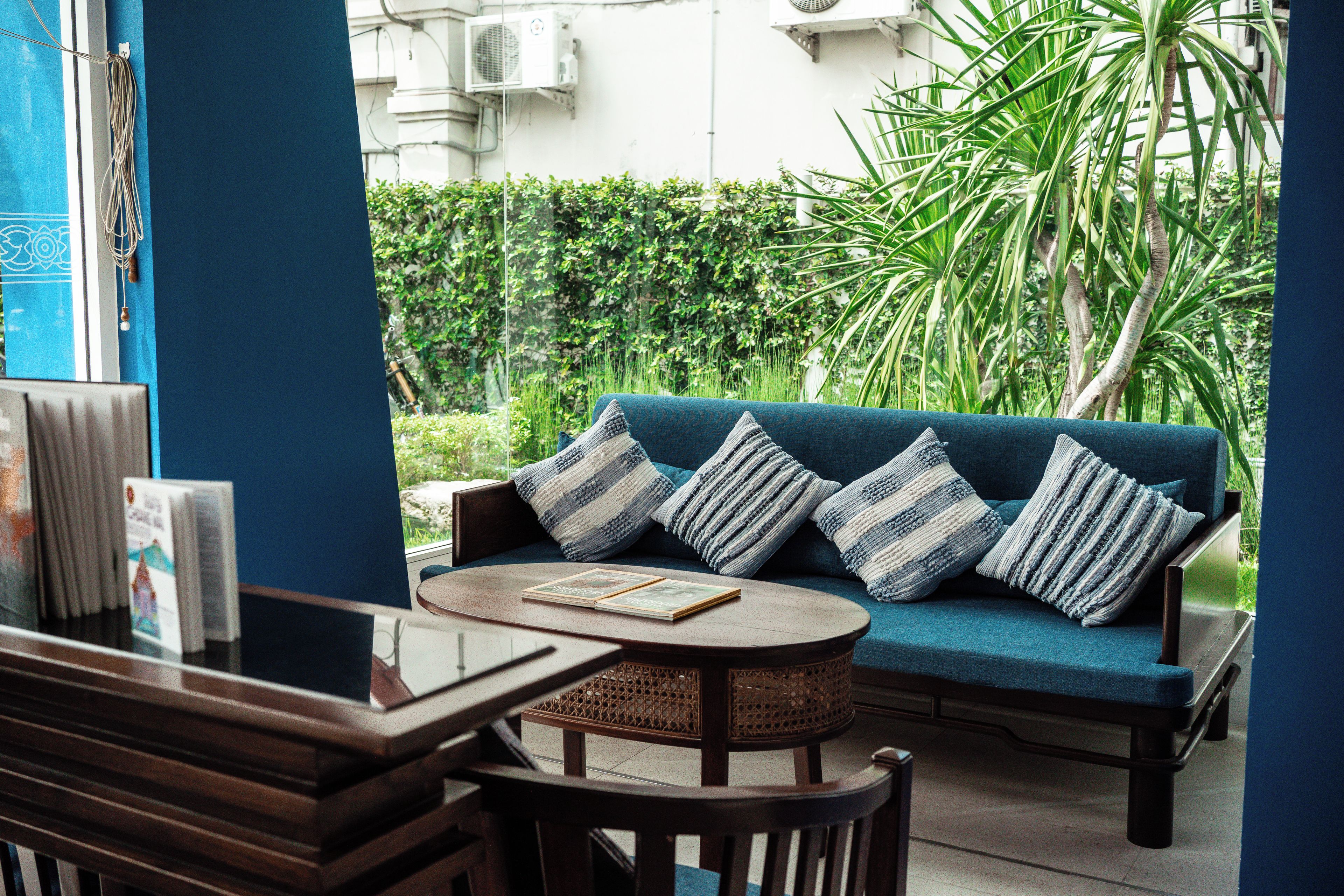 A blue sofa with striped cushions, a round wooden table, and a wicker chair in a room with a view of a garden and white building.
