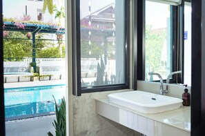 Une salle de bain avec un lavabo blanc, un miroir et une fenêtre donnant sur une piscine et un espace extérieur.