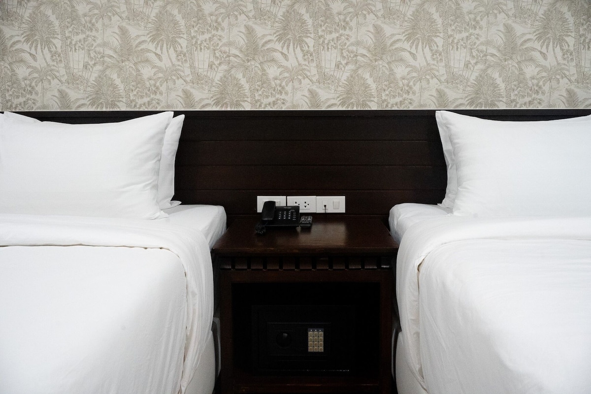 Een hotelkamer met twee bedden, een nachtkastje met telefoon en afstandsbediening, en een muur met een tropisch patroon.