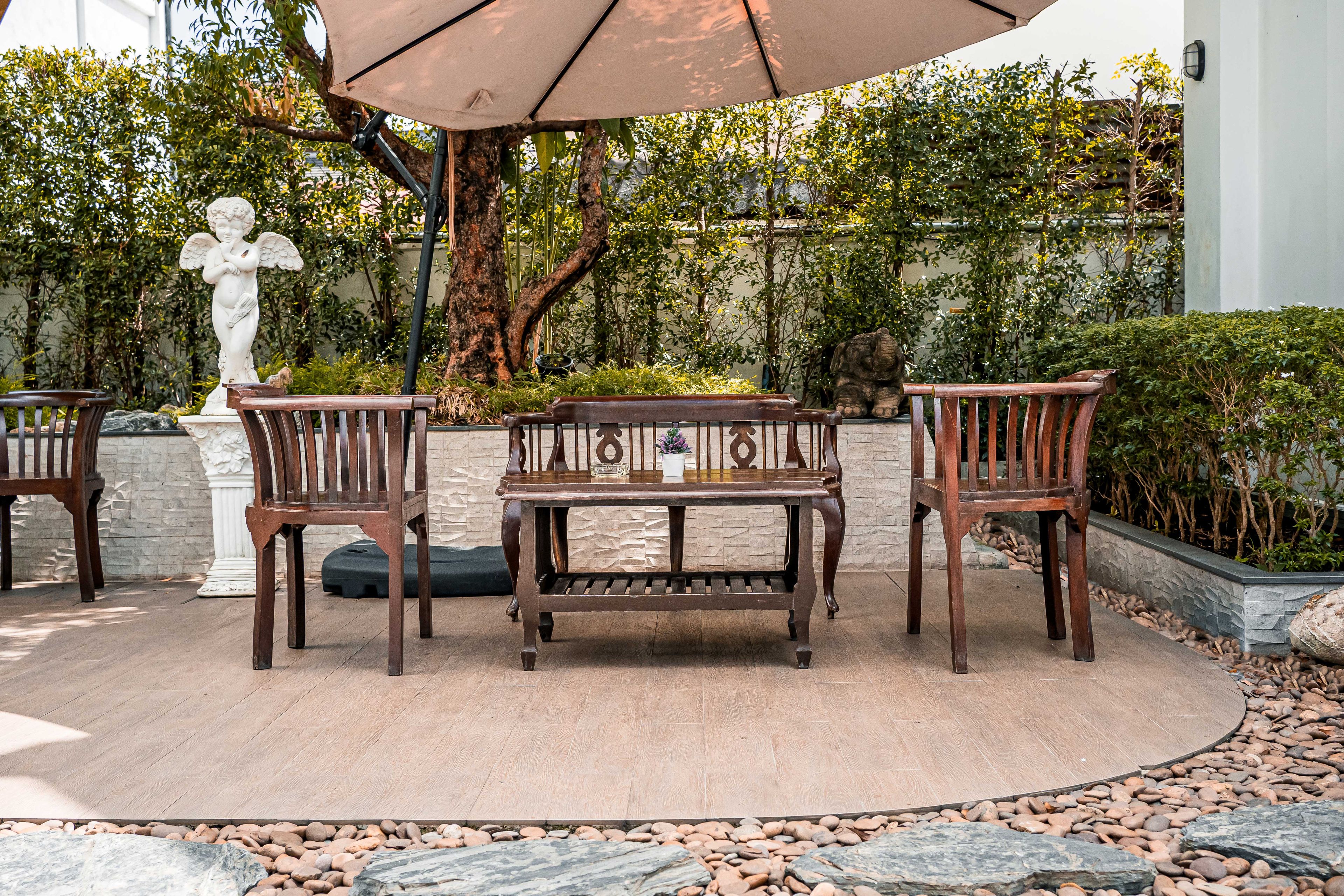 A wooden bench and table set under a large umbrella, surrounded by greenery and a statue.