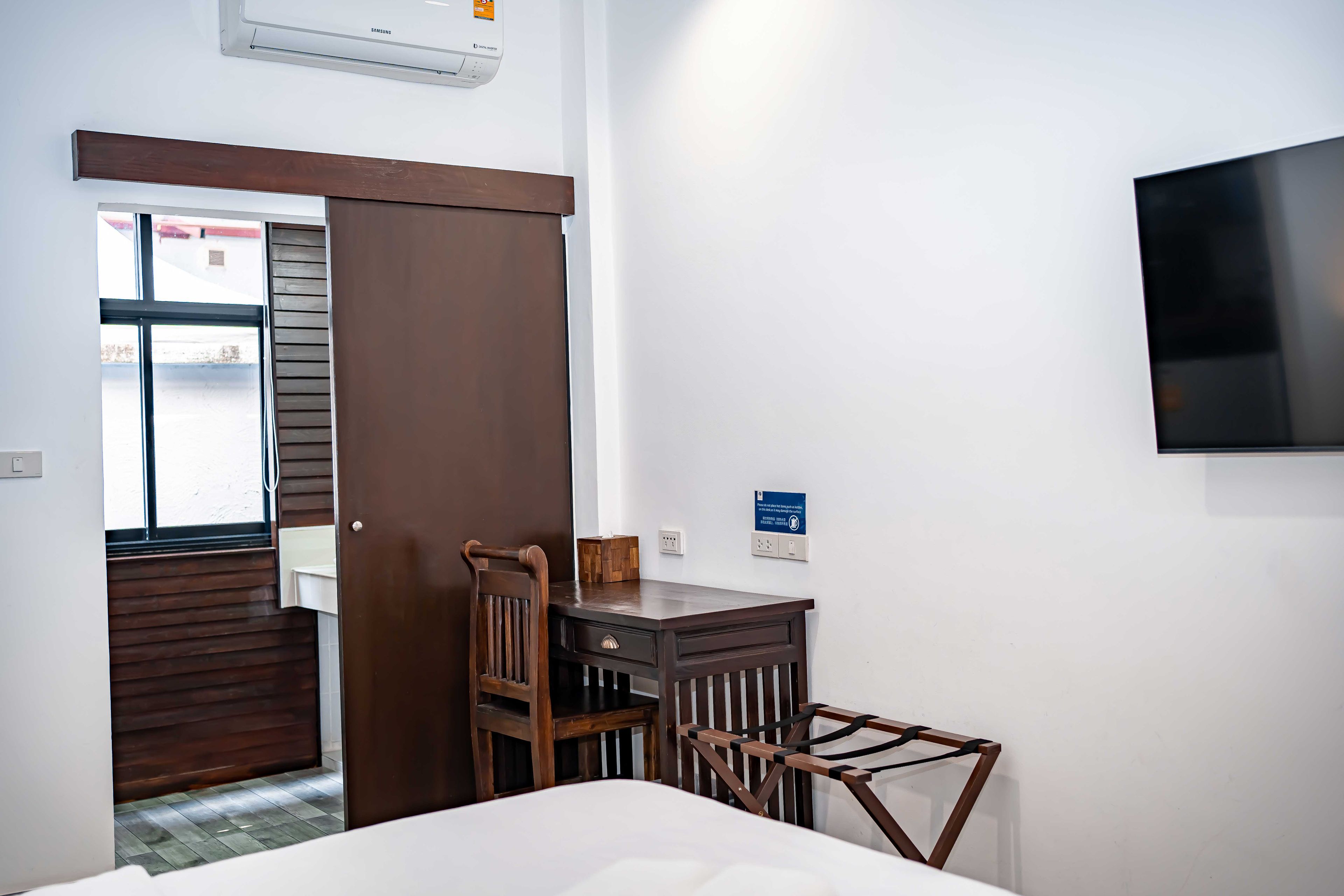A hotel room with a bed, a wooden chair, a small table, a TV mounted on the wall, and a window with blinds.