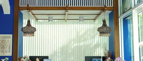 A reception desk with a wooden counter, two hanging lamps, a window with blinds, and a potted plant with flowers.